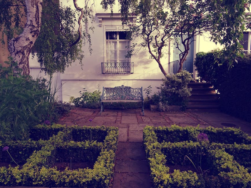 A small, well-maintained garden in front of a white house, featuring a paved patio area with a decorative metal bench positioned against the wall beneath a window with white shutters. The garden inclu