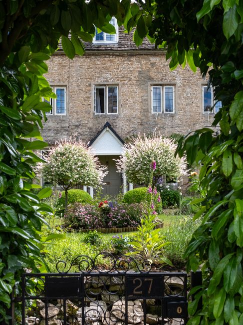 A small, well-maintained garden in front of a white house, featuring a paved patio area with a decorative metal bench positioned against the wall beneath a window with white shutters. The garden inclu