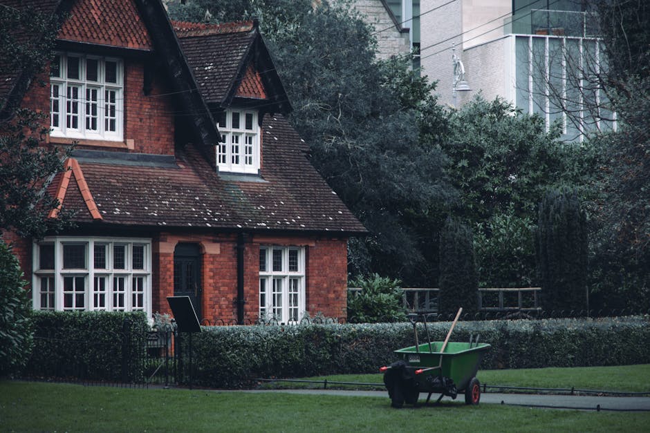 A red brick house with a steeply pitched roof and white-framed windows is surrounded by a well-maintained garden. The garden features a neatly trimmed hedge bordering the lawn, with lush green grass t